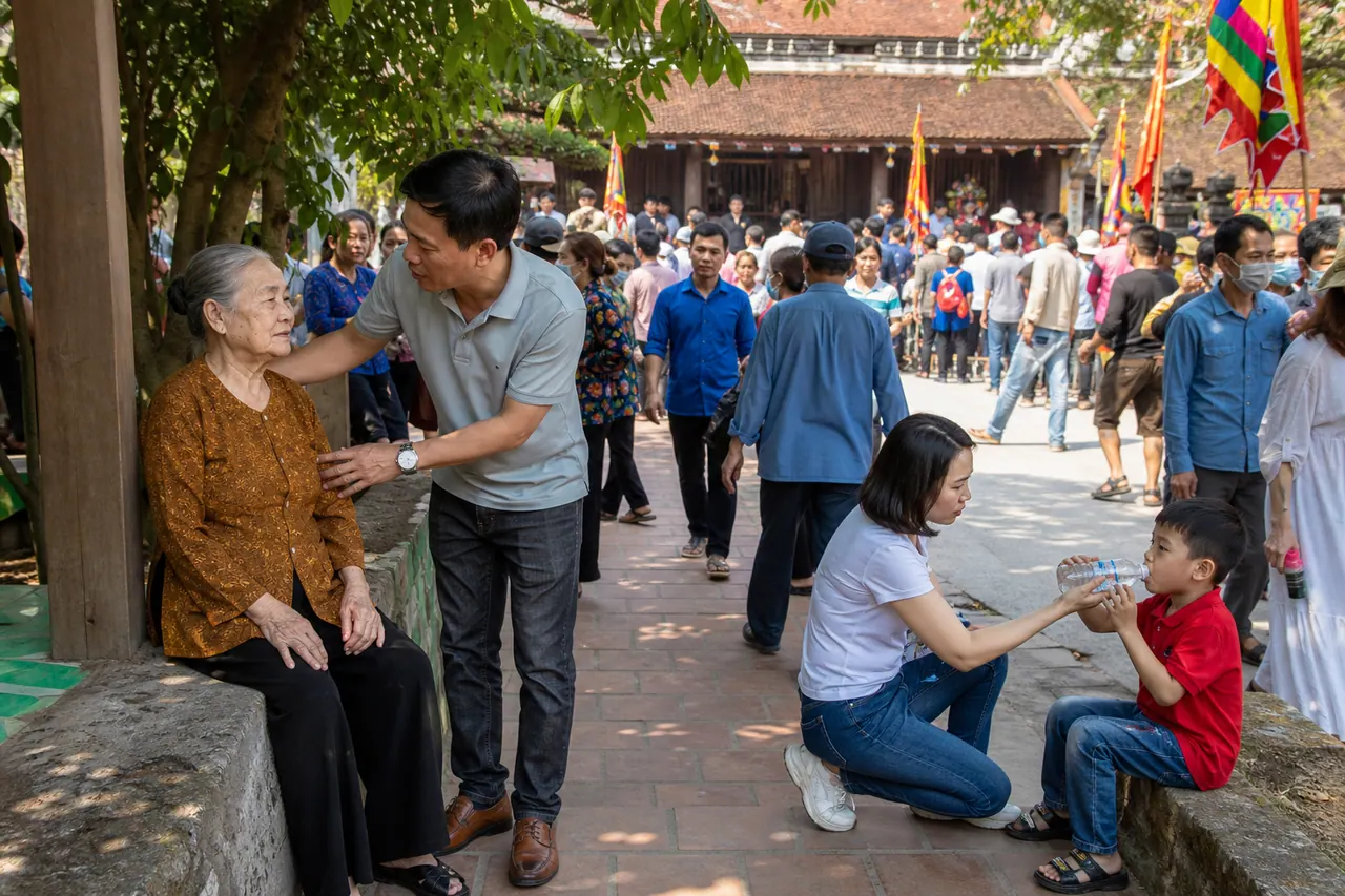 Người lớn đưa cụ già vào chỗ râm nghỉ chân và cho trẻ uống nước ở rìa khu hội đông