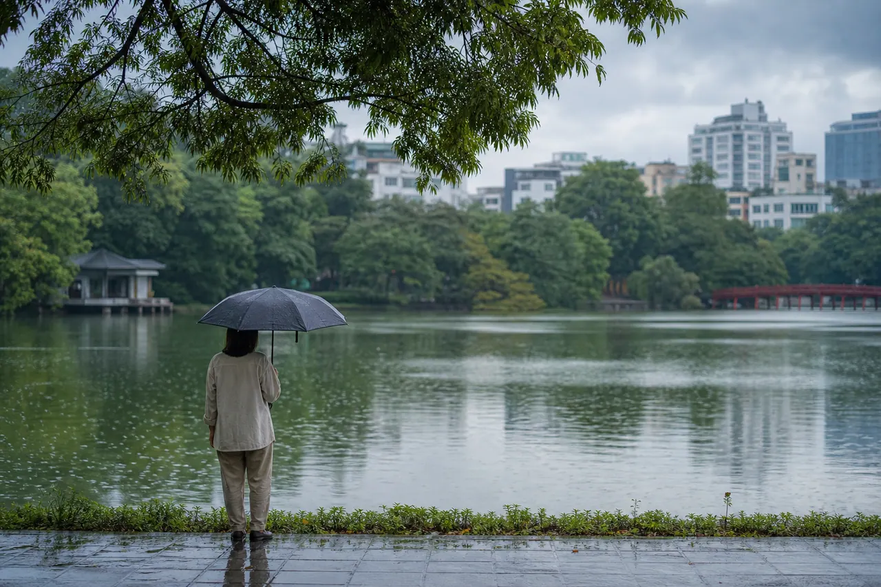 Người cầm ô đen đứng trên lối đi ven hồ, phía xa có công trình mái ngói và cầu đỏ