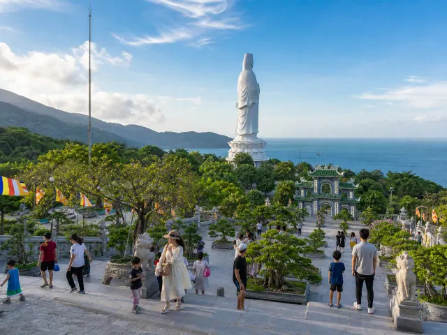 Tượng trắng quay lưng, cổng chùa, cây xanh và khách trên sân nhìn ra biển