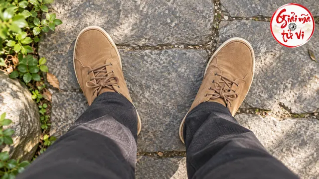 Hai chân mặc quần xám và giày nâu có mũi hướng ra hai bên trên nền đá, logo Giải Mã Tử Vi ở góc trên phải