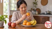 Người phụ nữ xem quả cam trước đĩa có cam và chuối, bên hai quả táo